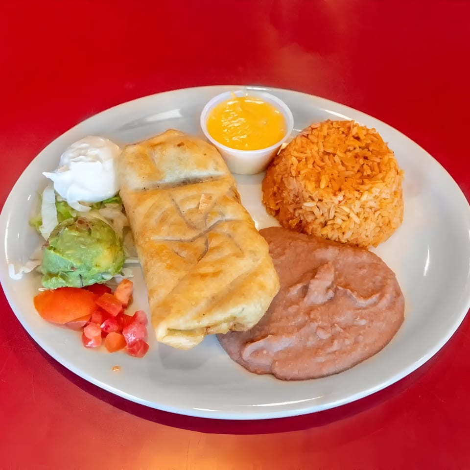 A fried burrito with rice, refried beans, guacamole, sour cream, and diced tomatoes on a white plate.