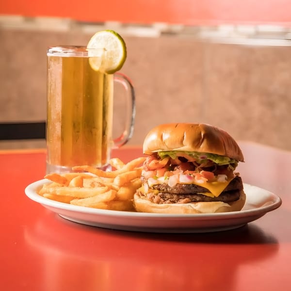 A double cheeseburger with pico de gallo, fries, and a beer with a lime on a red table.