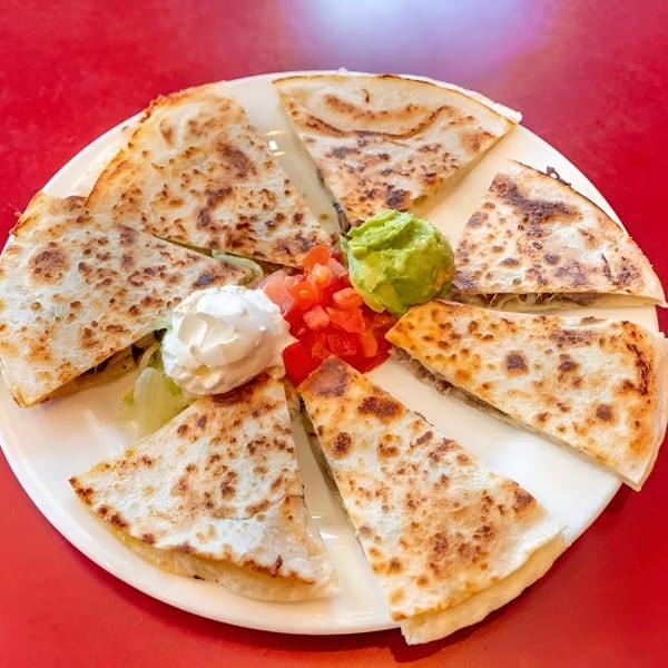 A white plate of quesadilla slices with guacamole, diced tomatoes, and sour cream on a red table.