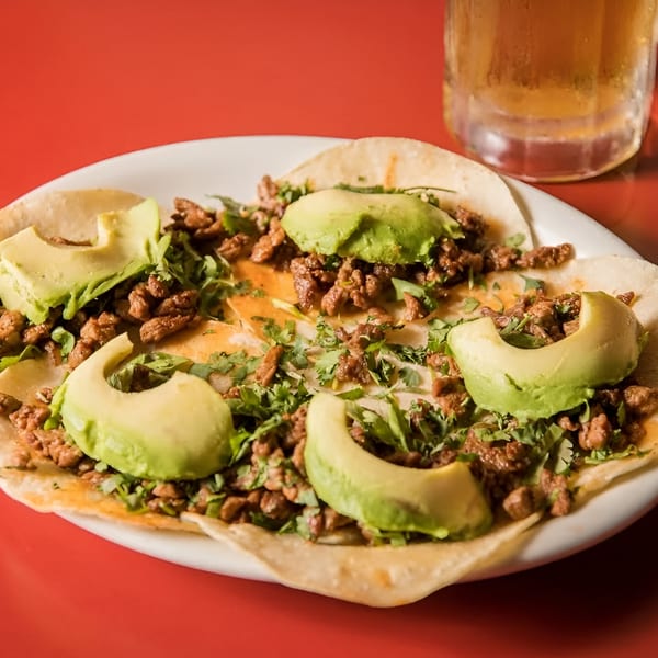 Four beef tacos with cilantro and avocado slices on a white plate with a beer.